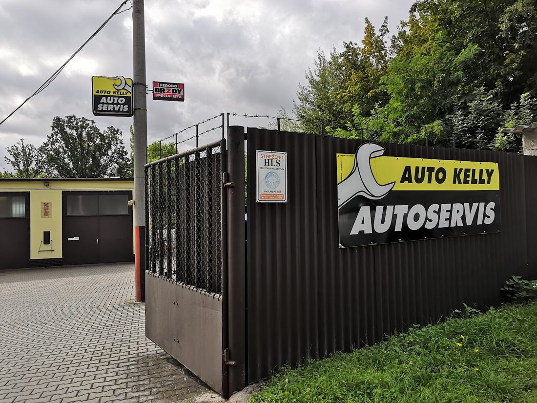 Tmavá plechová brána se žlutočernou cedulí Autoservis, za ní dvůr s garážemi